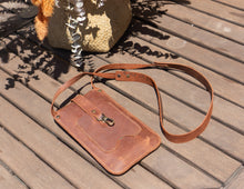 Alt Text: Handmade leather phone crossbody bag with a decorative background. Description: A handmade leather phone crossbody bag placed on a wooden table with a decorative background. The bag is brown with a front flap secured by a brass button and a long strap. The background includes flowers and a wicker basket.