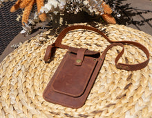 Alt Text: Handmade leather phone crossbody bag on a wicker surface. Description: A handmade leather phone crossbody bag lying on a woven wicker surface. The bag is brown with a front flap secured by a brass button and a long strap. The background includes part of a wicker basket and decorative flowers.
