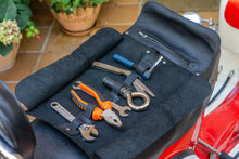 Open black leather motorcycle tool roll laid out on the seat of a red motorcycle, displaying various tools including a wrench, pliers, and a screwdriver. The roll features organized slots and a zippered pocket for secure storage.