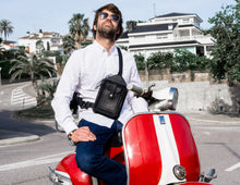 Man wearing a white shirt and blue jeans, with a black leather crossbody bag featuring detailed stitching and multiple pockets, sitting on a red and white scooter