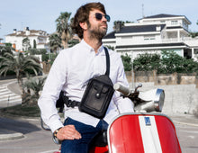 Man wearing a white shirt and blue jeans, sitting on a red and white scooter, showcasing a men's black leather crossbody bag with detailed stitching and multiple pockets.