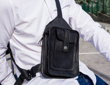 Close-up of a man wearing a white shirt and blue jeans, showcasing a men's black leather crossbody bag with detailed stitching and multiple pockets.