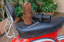 Brown and black leather motorcycle tool rolls, both rolled up and fastened with buckles, displayed on the seat of a red motorcycle, alongside a wrench.