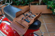 Open brown leather motorcycle tool roll displayed on the seat of a red motorcycle, featuring various tools and a zippered pocket for additional storage.