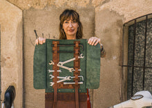 Handcrafted green and brown leather motorcycle tool roll opened with wrenches, held by woman in red pants, vintage scooter background, biker tool organizer