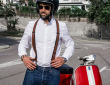 a front view of a person wearing brown reinforced leather suspenders over a white shirt, paired with blue jeans. They are standing next to a red and white scooter, wearing a helmet and sunglasses, ready to ride. The suspenders add a stylish, sophisticated touch to the outfit, with the leather intersecting at the back in a patch embossed with "Fashion Racing Handmade Custom Leather." The setting is an outdoor urban area with trees and buildings in the background.