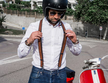 The image shows a person adjusting their brown reinforced leather suspenders over a white shirt, paired with blue jeans. They are standing next to a red and white scooter, wearing a helmet and sunglasses, ready for a ride. The suspenders intersect at a leather patch in the center of the back, which is embossed with a logo that reads "Fashion Racing Handmade Custom Leather." The setting is an outdoor urban area with trees and buildings in the background, along with a winding road.
