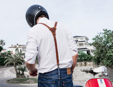 Back view of a man wearing a white shirt, blue jeans, and brown leather suspenders, standing next to a red and white scooter while wearing a black helmet