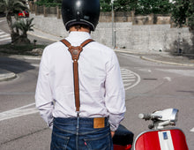 The image shows the back view of a person wearing brown black Reinforced leather suspenders over a white shirt, paired with blue jeans. They are sitting on a red and white scooter, with a helmet on, ready to ride. The suspenders intersect at a leather patch in the center of the back, which is embossed with a logo "Fashion Racing Handmade Custom Leather". The surroundings suggest an outdoor urban setting with trees and buildings in the background.