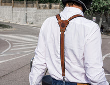 a back view of a person wearing brown reinforced leather suspenders over a white shirt, paired with blue jeans. They are sitting on a red and white scooter, wearing a helmet, and ready to ride. The suspenders intersect at a leather patch in the center of the back, which is embossed with a logo that reads "Fashion Racing Handmade Custom Leather." The surroundings depict an outdoor urban setting with trees and buildings in the background, along with a winding road.