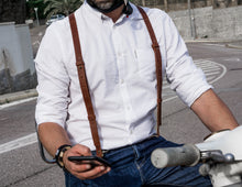 person wearing premium leather suspenders over a white button-down shirt. They are holding a smartphone and appear to be sitting on a motorcycle. The suspenders are brown and add a classic touch to the casual yet stylish outfit.