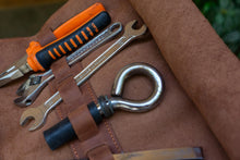 Close-up of an open brown leather motorcycle tool roll with organized tools including a wrench, pliers, and a hook, showcasing the tool slots and secure storage.