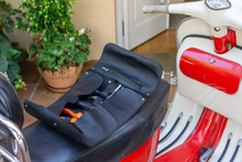 Open black leather motorcycle tool roll laid out on the seat of a red motorcycle, displaying various tools including a wrench, pliers, and a screwdriver. The roll features organized slots and a zippered pocket for secure storage.
