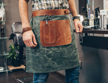 The image shows a person wearing a handmade waxed canvas leather apron. The apron is green with brown leather accents, particularly around the large front pocket and the belt area. The front pocket is holding a pair of scissors.