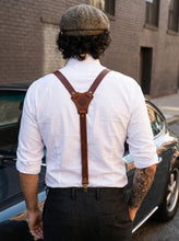 Men wearing a white shirt, brown suspenders, and a cap, standing in an urban setting.