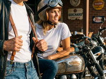 A woman in a vintage-style cream helmet with goggles sits on a chrome motorcycle next to a man in a leather jacket and white t-shirt, with a brick wall and vintage signs in the background.