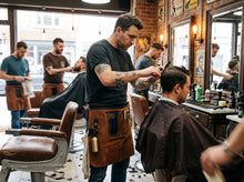 A tattooed barber wearing a brown leather apron剪s the hair of a customer seated in a classic barbershop with brick walls, mirrors, and other clients and staff in the background.