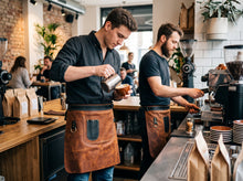Two baristas wearing brown leather aprons work behind a coffee counter in a modern cafe with customers and coffee equipment visible.
