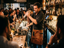 A bartender in a dark shirt and brown leather apron shakers a cocktail behind a busy bar counter with shelves of bottles and glasses.