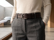 A close-up of a person wearing a dark brown embossed leather belt with a silver buckle, paired with gray trousers and a cream ribbed sweater.