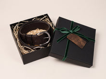 A dark brown leather belt with a silver buckle, presented inside an open black gift box with shredded paper filler. The closed black gift box with a green ribbon and a bronze 'Merry Christmas' tag sits beside it on a light beige background.