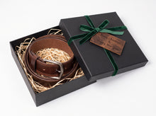 A brown leather belt with a silver buckle presented in an open black gift box filled with shredded paper, with the lid decorated in a dark green velvet ribbon and a wooden tag reading 'Merry Christmas & Happy New Year'.