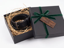 A black leather belt with a silver buckle presented in an open black gift box filled with straw, alongside the closed lid tied with a green velvet ribbon and a wooden tag that reads 'Merry Christmas & Happy New Year'.