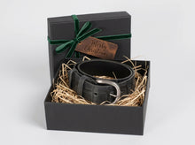A black leather belt with a silver buckle presented in a black gift box filled with straw, with the lid decorated in green velvet ribbon and a 'Merry Christmas' tag, on a light gray background.