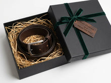 A dark brown leather belt with a silver buckle, presented in an open black gift box filled with straw, alongside the closed lid which is tied with a dark green ribbon and has a wooden tag reading 'Merry Christmas and Happy New Year!'.