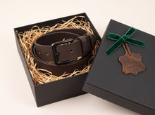 A dark brown leather belt with a black buckle, presented inside an open black gift box filled with shredded paper. The closed lid beside it has a green ribbon and a hanging tag that reads 'Merry Christmas'.