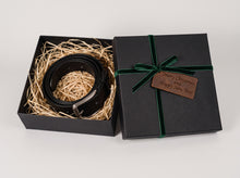 A black leather belt presented in a black gift box with shredded paper filler, alongside the matching lid wrapped in a dark green ribbon with a brown engraved tag reading 'Merry Christmas and Happy New Year'.