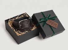 A dark brown leather belt with a silver buckle, presented inside an open black gift box with shredded paper filler. The matching black gift box lid sits beside it, tied with a dark green ribbon and featuring a small wooden tag that reads 'Thank you for your style and patience always.