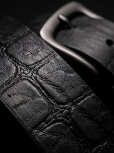 Close-up of a black leather watch strap with a crocodile-embossed texture and a dark metal buckle, photographed against a black background.