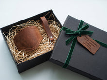 A brownleather motorcycle shifter boot protector with keichain presented in an open black gift box filled with shredded paper, next to a closed black gift box tied with a green ribbon and a brown tag 