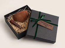 Brown leather motorcycle shifter boot protector with keichain presented in an open black gift box filled with shredded paper, next to a closed black gift box tied with a green ribbon and a brown tag that reads 'Happy Anniversary, My love, Your wife'.