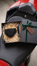 A black leather motorcycle boot protector presented in an open black gift box with shredded paper and a green ribbon, placed on a red and black Ducati motorcycle seat.