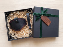 A black leather motorcycle boot protector with red stitching and a brass snap, presented in an open black gift box filled with shredded paper. The closed black gift box with a green velvet ribbon and a brown leather tag reading 'Happy Anniversary, my love' sits next to it on a light wood background.
