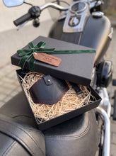 Gift box containing a black leather motorcycle boot protector with red contrast stitching and a matching keychain, presented on natural wood wool with a green velvet ribbon and engraved gift tag, displayed on the seat of a black cruiser-style motorcycle.