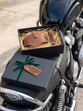Brown leather motorcycle shifter boot protector and matching leather keychain gift set in a premium gift box on a Triumph motorcycle seat, handcrafted leather motorcycle boot protector designed to prevent shifter wear and protect riding boots.