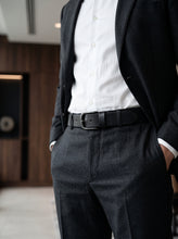 A close-up of a person wearing a dark charcoal gray suit with a white button-down shirt, a black leather belt with a silver buckle, and a black blazer, standing in a modern indoor setting with a blurred wooden background.