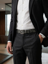 A man wearing a dark charcoal gray suit with a white dress shirt and a dark gray textured leather belt with a silver buckle, standing in a modern office environment.