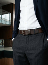A close-up of a person wearing a dark navy blazer, white dress shirt, dark gray trousers, and a brown leather belt with a silver buckle in an indoor setting.
