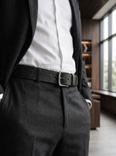 A close-up of a man wearing black leather belt croc pattern with a silver rectangular buckle, styled with a white dress shirt and dark suit jacket and trousers in an indoor setting.