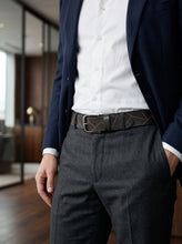 A man wearing a dark brown geometric-patterned leather belt with a silver buckle, styled with a white dress shirt, navy blazer, and dark gray trousers.