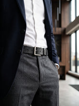 A close-up of a man wearing a dark navy blazer, white dress shirt, charcoal gray trousers, and a black leather belt with a silver buckle in a modern indoor setting.