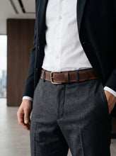 A close-up of a man wearing a dark navy blazer, white dress shirt, brown leather belt with a silver buckle, and dark gray textured trousers, standing in a modern office setting.