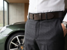 A close-up of a man wearing a dark brown leather belt with a silver buckle, styled with a white dress shirt and dark gray trousers. A green sports car is blurred in the background.