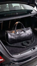 A black leather duffel bag with dual top handles and a detachable shoulder strap, displayed inside the open trunk of a black Mercedes-Benz car.