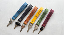 Six personalized leather keychains in different colors (blue, black, brown, yellow, green, and purple) with engraved names and phone numbers, each attached to a metal key ring and key, arranged on a light gray background.