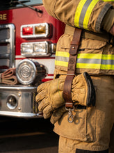 Brown Leather Glove Strap Holder attached to a firefighter turnout belt and securely holding heavy-duty firefighter gloves in front of a fire truck. Handmade genuine leather glove holder with an adjustable belt loop, durable leather strap, and heavy-duty swivel snap hook for carrying firefighter gloves, rescue gloves, work gloves, and protective gear hands-free. Personalized leather glove strap holder, firefighter gear accessory, practical EDC tool, and thoughtful gift for firefighters and first responders.